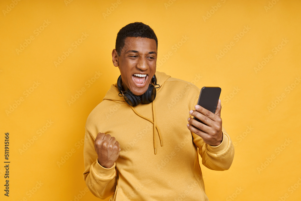 Excited mixed race man with headphones using smart phone while standing against yellow Excited mixed race man with headphones using smart phone while standing against yellow