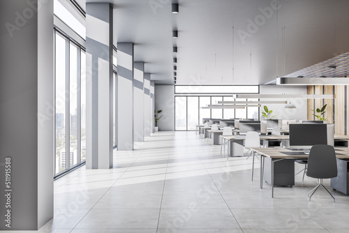 Spacious sunlit coworking office with wooden work tables, concrete floor, grey stylish lamps on light ceiling and city view from panoramic windows. 3D rendering