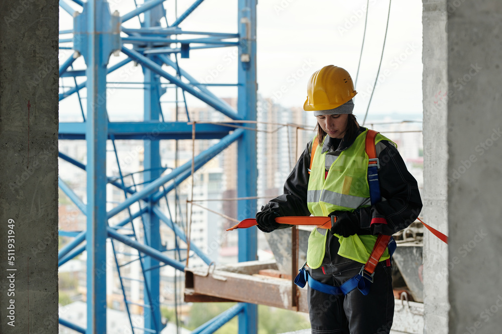 Young woman in protective helmet and reflective vest fastening safety belt on her waist while going to work at construction site Young woman in protective helmet and reflective vest fastening safety belt on her waist while going to work at construction site