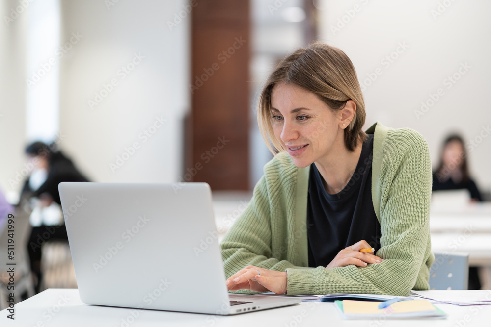 Smiling middle-aged woman studying online on laptop computer while sitting in classroom, doing second degree as mature student. Happy 45s female attending online professional development course Smiling middle-aged woman studying online on laptop computer while sitting in classroom, doing second degree as mature student. Happy 45s female attending online professional development course