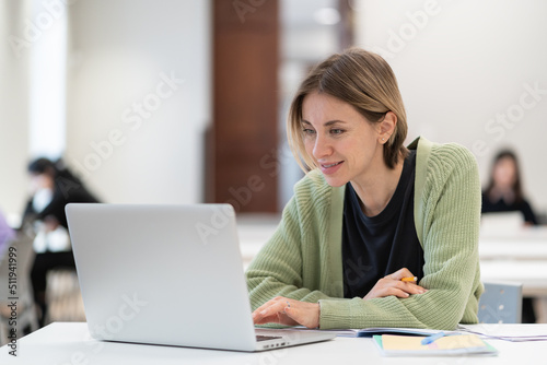 Smiling middle-aged woman studying online on laptop computer while sitting in classroom  doing second degree as mature student. Happy 45s female attending online professional development course