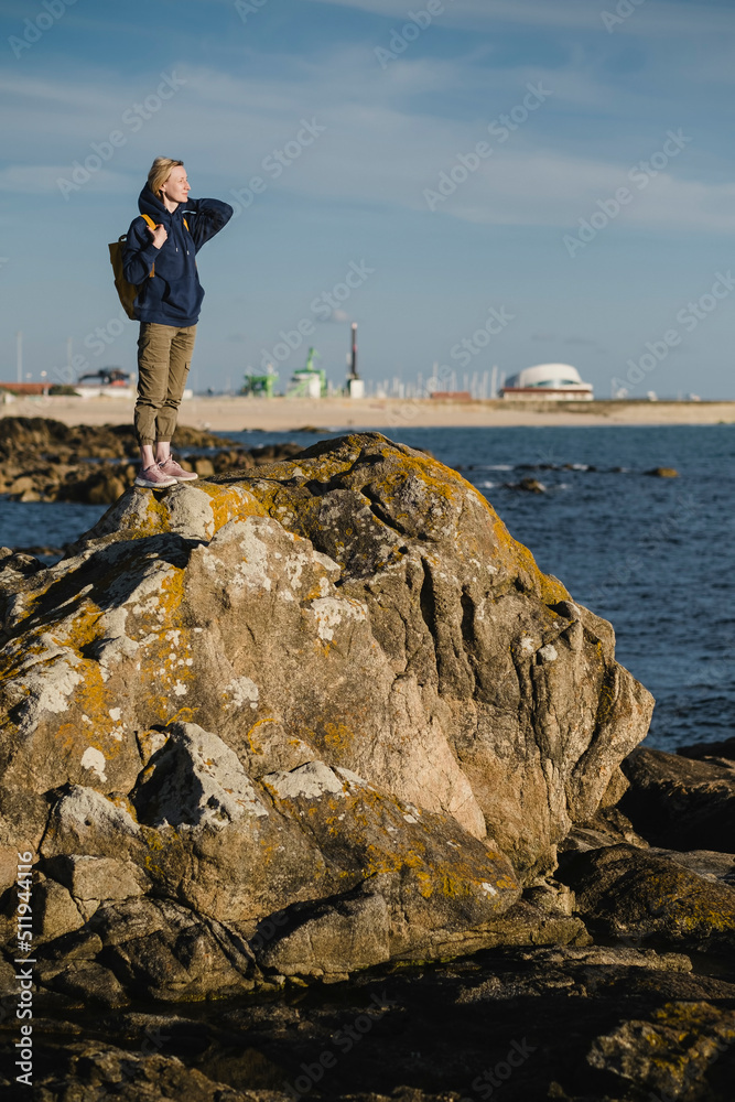 A female with a yellow backpack stands on the coastal ocean rocks.