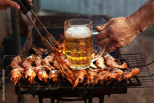 large shelled langoustine shrimp lie on a grill with smoke. Barbecue outdoors, human hands prepare food on a fire and hold beer in a glass photo