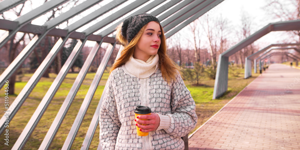 Modern teenage hipster girl in spring in park in hat with bubo and knitted sweater, with takeaway coffee