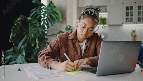 Young focused african american woman employee writing note and marking documents, working online on laptop, zoom out