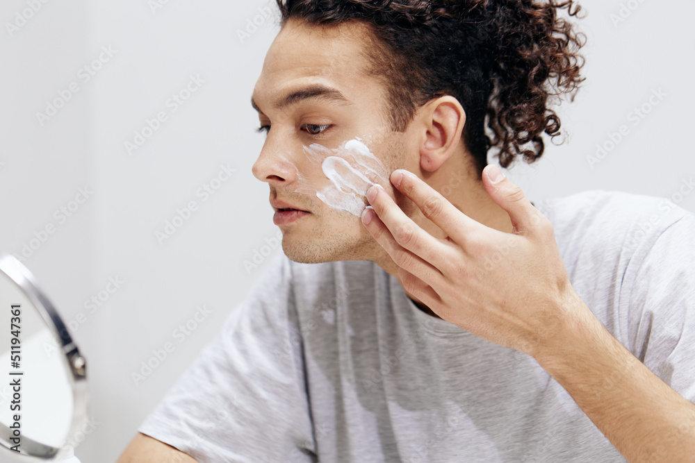 A young man in a white T-shirt in front of a mirror skin care light background