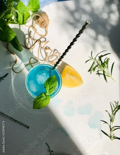 Blue cold drink in a glass with ice cubes on white desk with blue background. Also used mint leaves and rosemary, rope, spoon with natual sunlight from the open window
