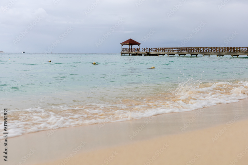 A pier goes into the sea,at the end there is a gazebo.The blue sea water and the sky are separated by the horizon line,a pier.Caribbean Coast in the Dominican Republic.A wave runs over the sandy shore