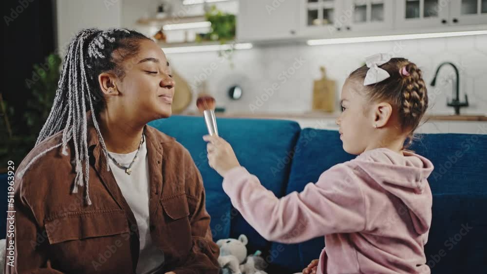 Playful little african american girl doing make up for her pretty mommy, applying rouge on her face with big brush Playful little african american girl doing make up for her pretty mommy, applying rouge on her face with big brush