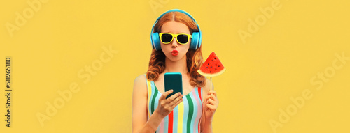 Summer colorful portrait of stylish young woman in headphones listening to music on smartphone with juicy lollipop or ice cream shaped slice of watermelon on blue background © rohappy