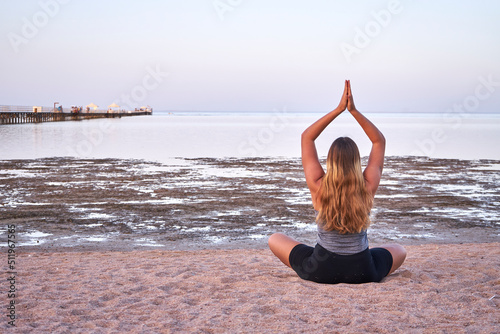 Young long hair woman practicing yoga on the beach at sunset. Caucasian girl with long blond hair doing yoga exercises on a sandy beach at sunrise.   
