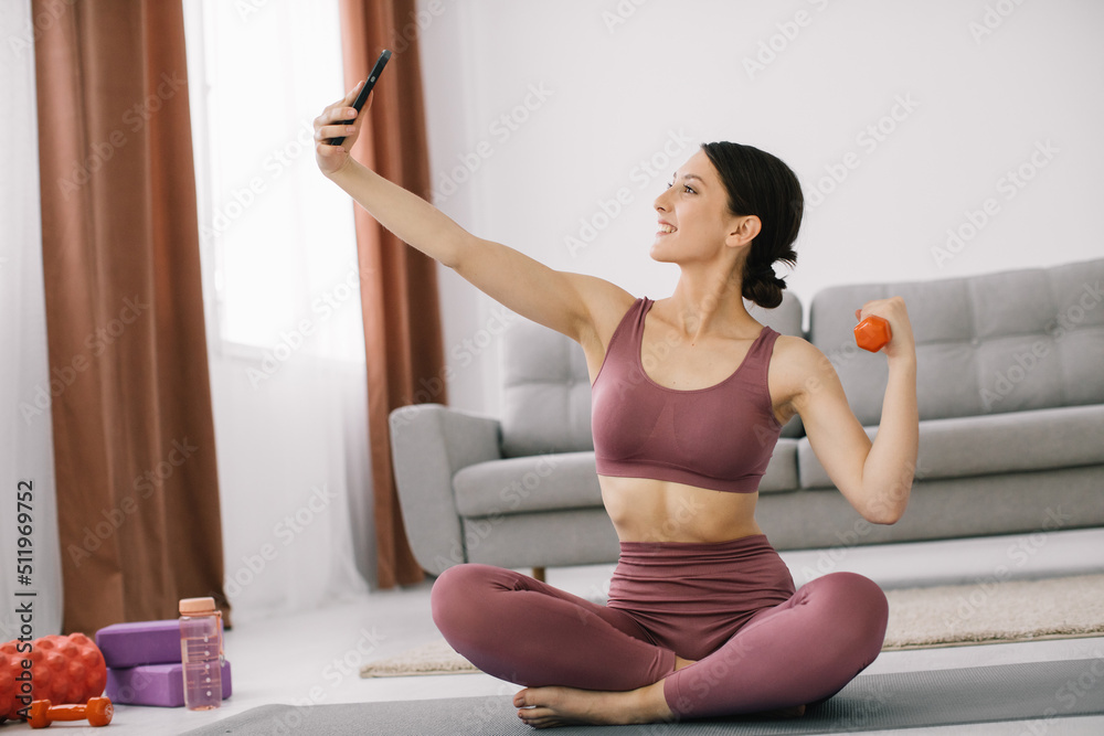Attractive young woman taking selfie showing biceps with dumbbell at home.