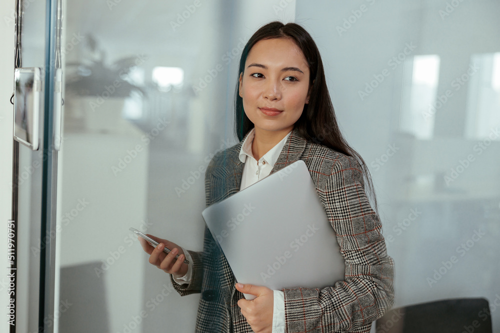 Charming Asian business woman with a smile standing holding laptop and mobile phone at the office Charming Asian business woman with a smile standing holding laptop and mobile phone at the office
