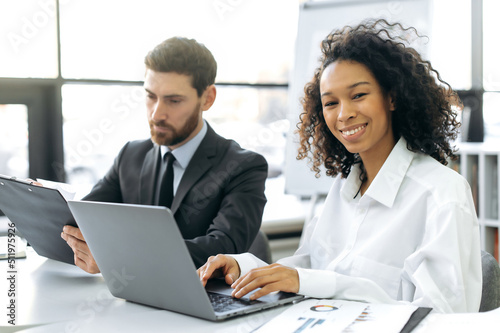 International colleagues working in a modern office, caucasian man, manager, working with documents, African American woman, financial consultant, working at a laptop, looking at the camera, smiling