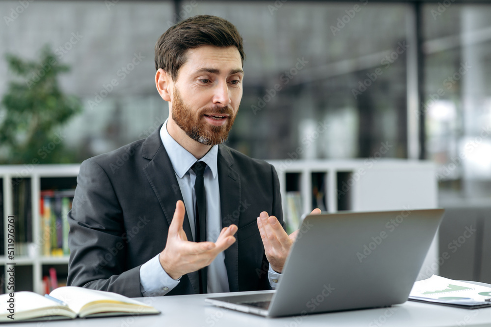 Busy attractive Caucasian businessman, company owner, insurance manager, sits in a modern office at work desk, holds a video call with a client, online consultation, gesturing hands, smiling friendly