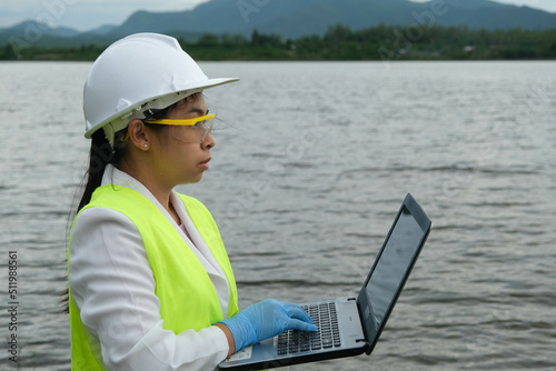 Female environmentalist wearing gloves takes samples of river water to examine contaminants in natural waters and record data on a laptop. Water and ecology concept. © Pornpimon