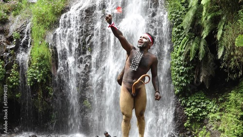 Papua man wearing traditional clothes of Dani tribe, red-white headband and bangle is holding little Indonesia flag and celebrating Indonesia independence day against waterfall background photo