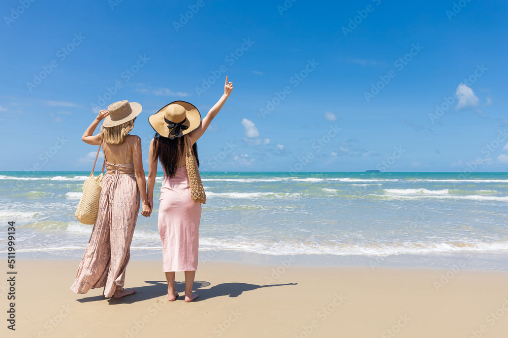 back view women couple standing on the beach and pointing to the sky during summer vacation