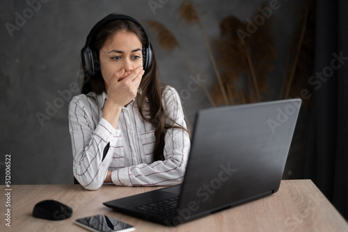 Portrait of shocked young woman watching horror film at home using headphone and laptop. She is covering her mouth by hand with surprise while staring at a computer