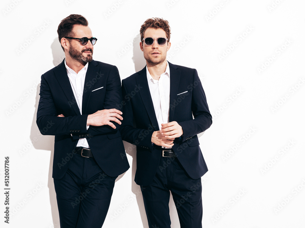 Portrait of two handsome confident stylish hipster lambersexual models. Sexy modern men dressed in black elegant suit. Fashion male posing in studio near white wall in sunglasses Portrait of two handsome confident stylish hipster lambersexual models. Sexy modern men dressed in black elegant suit. Fashion male posing in studio near white wall in sunglasses