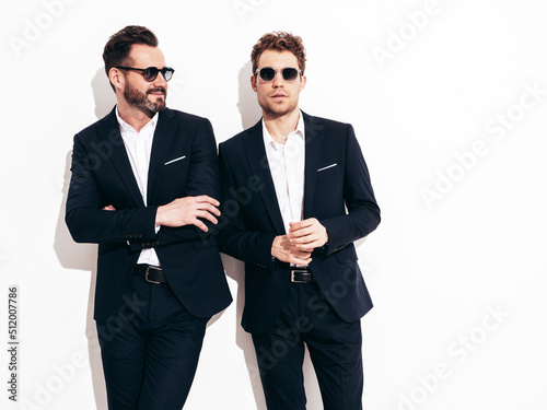 Portrait of two handsome confident stylish hipster lambersexual models. Sexy modern men dressed in black elegant suit. Fashion male posing in studio near white wall in sunglasses