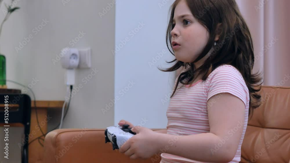 A little brunette girl with curly hair is playing video games sitting on the couch at home, upset or offended, focused, stuck out her tongue, says stretches her lips. A little brunette girl with curly hair is playing video games sitting on the couch at home, upset or offended, focused, stuck out her tongue, says stretches her lips.
