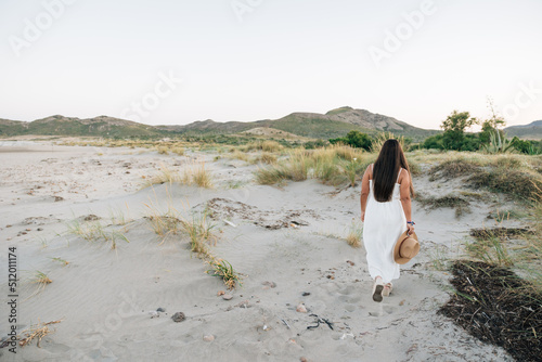 Unrecognizable woman wearing a white dress and holding a hat while walking outdoors in nature.