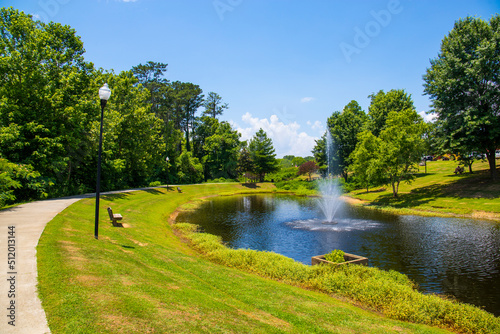 a gorgeous summer landscape in the park with a pond with a water fountain surrounded by lush green trees, grass and plants with blue sky and clouds at Logan Farm Park in Acworth Georgia USA photo