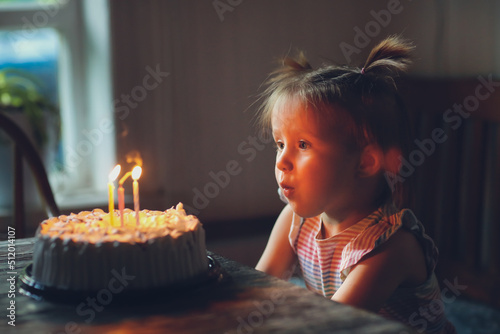 Funny toddler girl with two ponytails will conceive 3 candles on birthday cake, dark style and soft focus