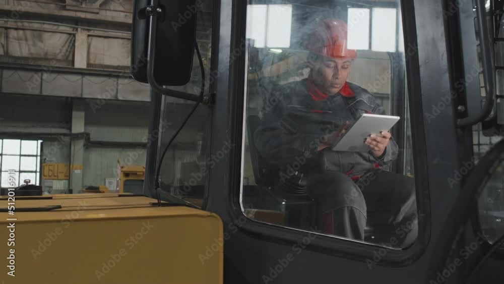 Medium long of Black woman in orange hard hat and work overalls sitting inside cabin of heavy machinery at factory, testing it, using tablet Medium long of Black woman in orange hard hat and work overalls sitting inside cabin of heavy machinery at factory, testing it, using tablet