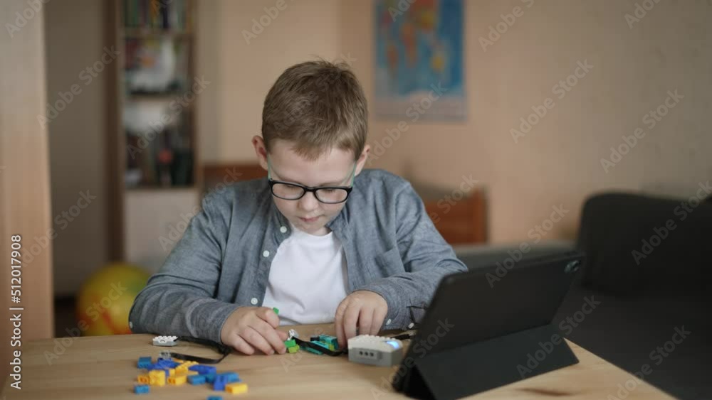 Caucasian boy with glasses creates his own model of programmable robot from plastic constructor. Child sits at table, clicks on screen in tablet and connects the details of the toy. Caucasian boy with glasses creates his own model of programmable robot from plastic constructor. Child sits at table, clicks on screen in tablet and connects the details of the toy.