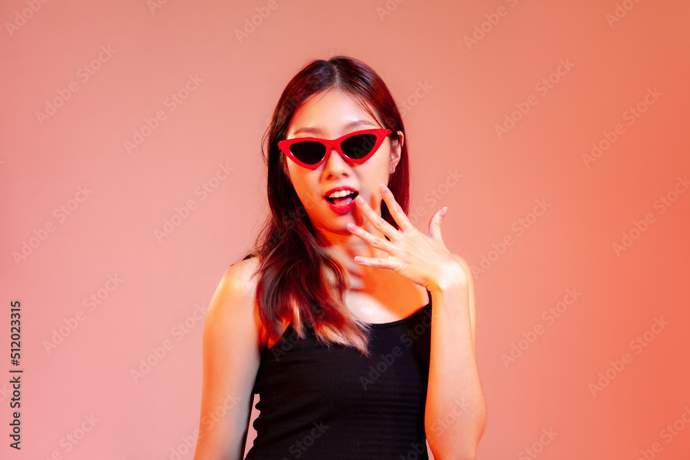 Fototapeta premium Young asian woman in black tank top wearing red sunglasses posing exciting and surprised on red pastel background.