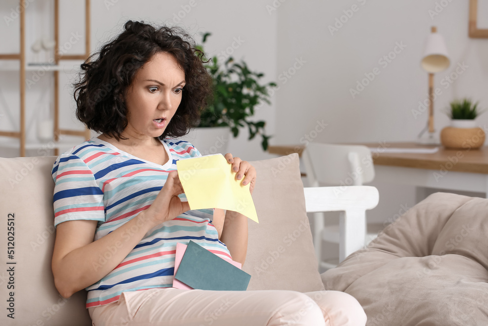 Shocked pregnant woman opening envelope on couch at home Shocked pregnant woman opening envelope on couch at home