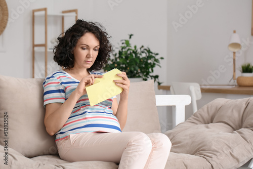 Young pregnant woman opening envelope on couch at home