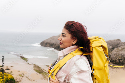 Smiling woman with red hair wearing backpack standing in front of sea