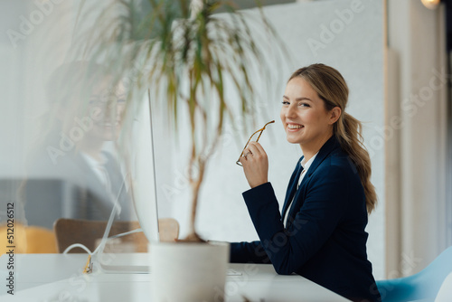 Smiling businesswoman holding eyeglasses sitting at desk in office