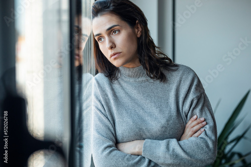 Depressed woman standing with arms crossed looking through window at home photo