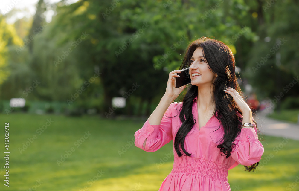Close up portrait of a young brunette with wavy hair dressed in a pink dress walking in the park talking on a cell phone. Close up portrait of a young brunette with wavy hair dressed in a pink dress walking in the park talking on a cell phone.