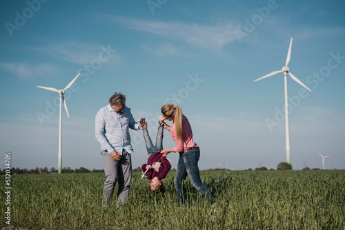 Playful parents carrying daughter upside down with wind turbines in background at wind farm