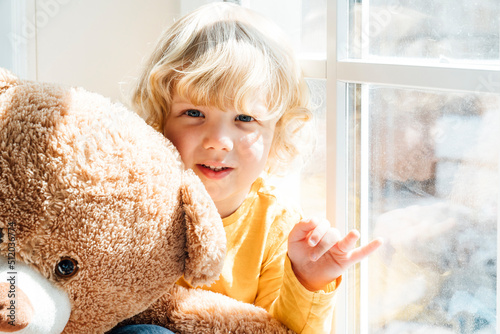 Cute boy with teddy bear sitting by window at home photo