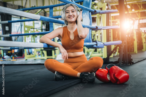 Cute boxing woman sitting and listening song with headphone relaxing in the gym. Smiling woman sitting on the floor after hard work out.