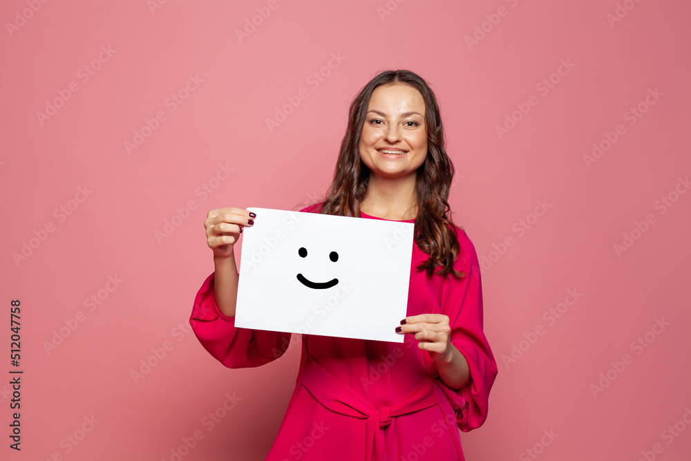 Young woman holding white paper with smile sign on pink background Young woman holding white paper with smile sign on pink background