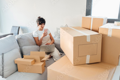 Woman using mobile phone sitting with laptop and cardboard boxes in living room photo