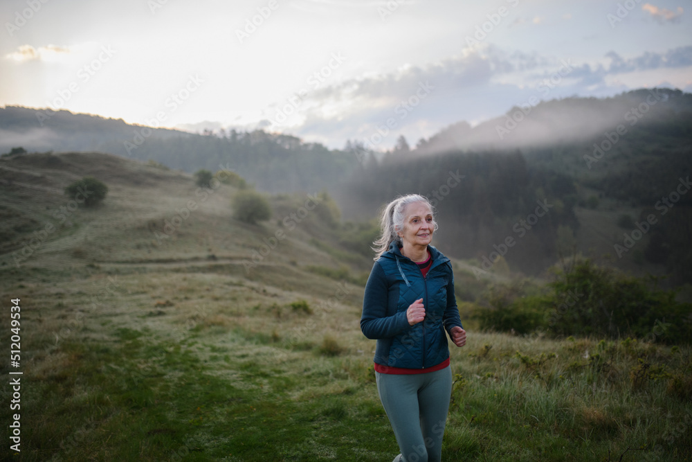 Fototapeta premium Senior woman jogging in nature on early morning with fog and mountains in background.
