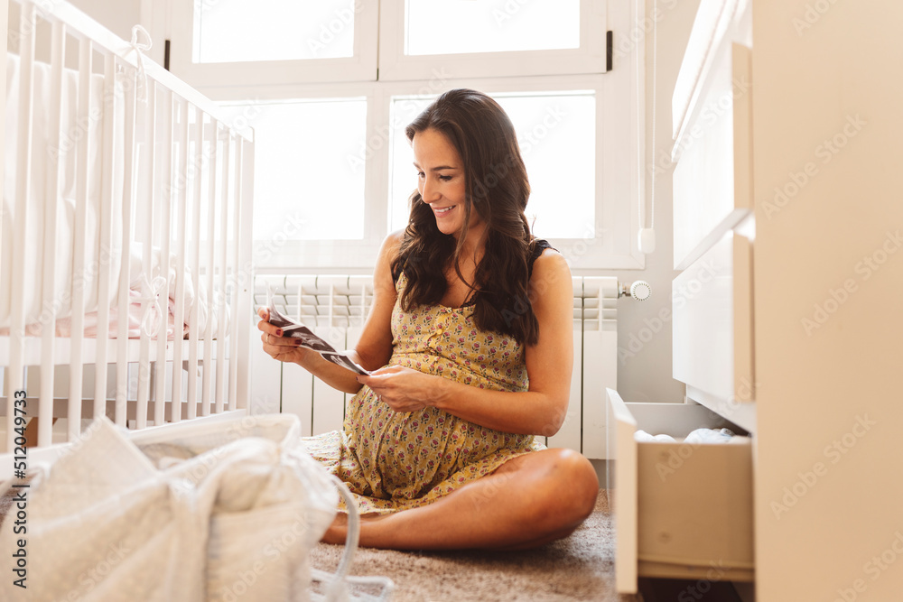 Happy pregnant woman with ultrasound scans sitting by crib at home Happy pregnant woman with ultrasound scans sitting by crib at home