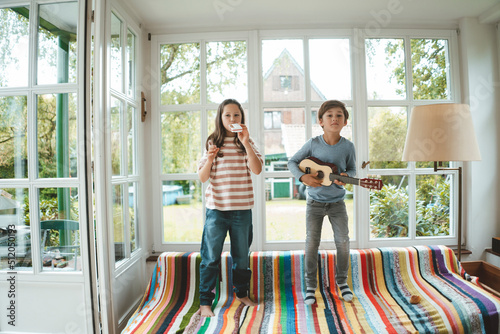 Siblings playing musical instrument standing on sofa at home photo