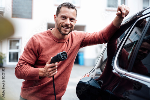 Smiling man holding charging cable leaning on electric car photo