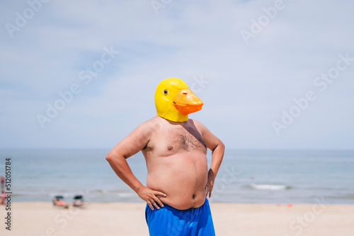 Shirtless obese man with hand on hips wearing duck mask standing at beach