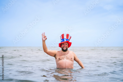 Smiling shirtless obese man wearing Uncle Sam hat showing OK sign in sea photo