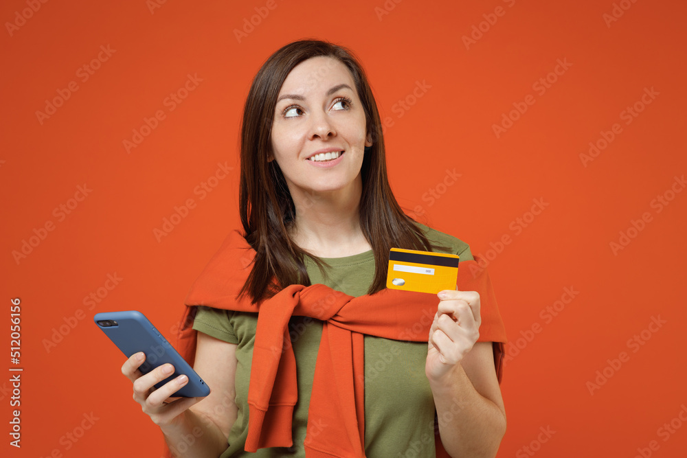 Young woman in khaki t-shirt tied sweater on shoulders using mobile cell phone hold credit bank card shopping online order delivery booking tour look aside on area isolated on plain orange background. Young woman in khaki t-shirt tied sweater on shoulders using mobile cell phone hold credit bank card shopping online order delivery booking tour look aside on area isolated on plain orange background.
